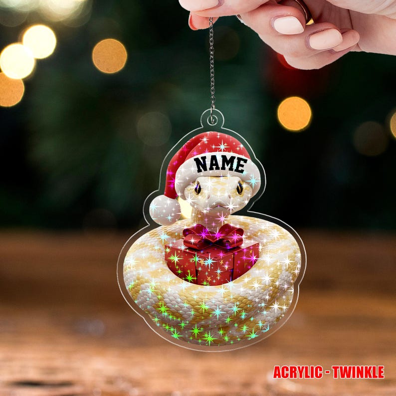 May include: A clear acrylic Christmas ornament featuring a snake wearing a Santa hat with the word "NAME" on it. The snake is coiled around a red gift box with a pink bow. The ornament has a glittery, star-like effect.