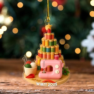 May include: A Christmas tree ornament featuring a sewing machine and spools of thread. The tree is made of colorful thread spools, with a pink sewing machine at the base. The ornament includes a basket of yarn, needles, and the text "WYATT 2025".