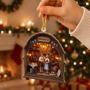 May include: A decorative hanging ornament shaped like a wooden archway with the text Oktoberfest. The interior depicts a detailed scene of two figures in traditional clothing holding beer mugs inside a tavern with shelves, barrels, and warm lighting.