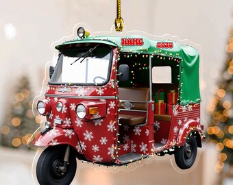 May include: Red and green Christmas ornament shaped like a tuk-tuk, decorated with snowflakes and string lights. The ornament has the word "NAME" and the year "2023" on it. The tuk-tuk has a brown seat and several wrapped gifts inside.
