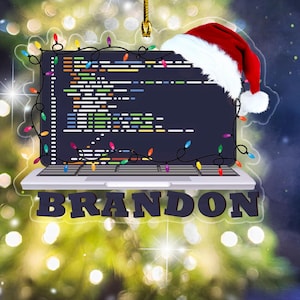 May include: A Christmas ornament shaped like a laptop displaying code, with a Santa hat on top. The ornament is decorated with colorful string lights and the name "BRANDON" in bold letters.