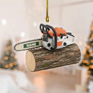May include: A chainsaw ornament with a white and orange body, black handles, and a silver chain, resting on a wooden log. The text "NAME" is on the chainsaw blade. The ornament hangs from a gold string.
