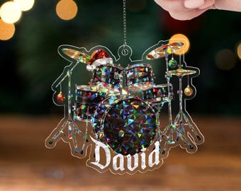 May include: A clear acrylic Christmas ornament shaped like a drum set with a Santa hat on top. The drum set is decorated with colorful geometric shapes and the name "David" is written in white script. The text "WAVY ACRYLIC - CRACKLE" is at the bottom.