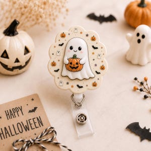 May include: A circular badge reel with a scalloped edge featuring a cream background decorated with small orange pumpkins, black bats, and stars. A central white ghost illustration holds a small orange jack-o'-lantern with a smiling face.
