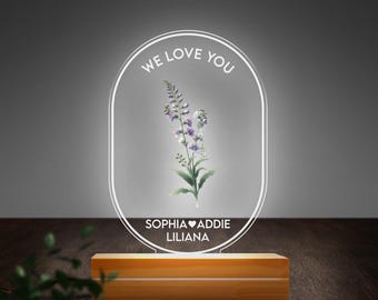 May include: A white oval-shaped acrylic sign with the text "We Love You" and a floral illustration. The sign is on a wooden base and has the names "Sophia, Addie, Liliana" with a heart symbol between the first two names.