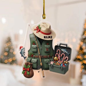 May include: A Christmas ornament featuring a fishing theme. The ornament includes a green fishing vest, a tan hat with a red bow, a fishing rod, a tackle box with candy canes, and a fish wearing a Santa hat. The word "NAME" is printed on the hat.