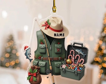 May include: A Christmas ornament featuring a fishing theme. The ornament includes a green fishing vest, a tan hat with a red bow, a fishing rod, a tackle box with candy canes, and a fish wearing a Santa hat. The word "NAME" is printed on the hat.