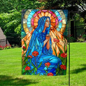 May include: A stained glass style garden flag featuring a woman in a blue robe with her hands clasped in prayer. The background is a yellow and orange stained glass design with red flowers.
