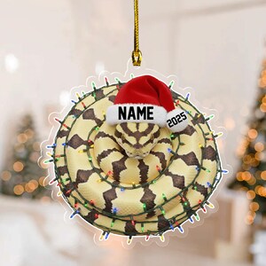 May include: A decorative ornament featuring a coiled snake with a Santa hat. The snake is tan with brown markings and is encircled by a string of colorful Christmas lights. The hat has the word "NAME" and the year "2025" printed on it.