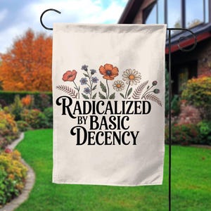 May include: A cream-colored garden flag with the text "RADICALIZED BY BASIC DECENCY" in black script. The flag is decorated with a floral design featuring orange poppies, blue wildflowers, and white daisies. The flag is hanging on a black metal stand.