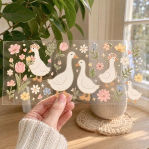 K&ouml;nnte beinhalten: Ein transparenter Stickerbogen mit wei&szlig;en G&auml;nsen mit orangefarbenen Schn&auml;beln und F&uuml;&szlig;en, durchsetzt mit verschiedenen bunten Blumen wie rosa Tulpen, gelben Bl&uuml;ten, kleinen blauen Bl&uuml;tenbl&auml;ttern sowie gr&uuml;nen Bl&auml;ttern und kleinen funkelnden Sternen.