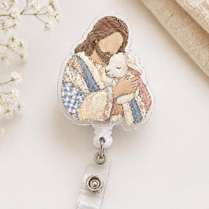 May include: A badge reel featuring a clear acrylic topper with a printed illustration of a figure with long brown hair and a beard holding a small white lamb. The figure wears a robe with a patchwork pattern of blue, pink, and cream floral fabrics.