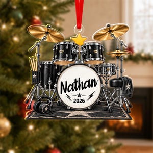 May include: A flat, decorative ornament shaped like a drum set with black drums, gold cymbals, and a white bass drum head featuring the name Nathan and the year 2026. The design includes a pair of drumsticks, headphones, and a red hanging ribbon.