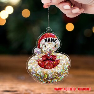 May include: A clear acrylic Christmas ornament shaped like a snowman wearing a Santa hat with the word "NAME". The snowman holds a red gift box. The ornament has a wavy acrylic crackle design. The text "WAVY ACRYLIC - CRACKLE" is at the bottom.