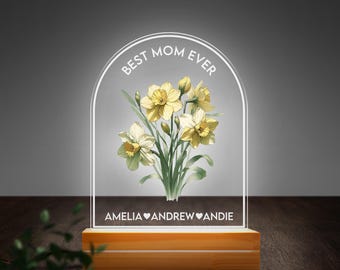 May include: A white acrylic lamp with a floral design and the text "Best Mom Ever". The base of the lamp is made of wood and has the names "Amelia, Andrew, and Andie" inscribed on it.