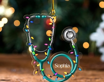 May include: A teal stethoscope Christmas ornament with the name "Sophia" printed on it. The ornament is decorated with colorful Christmas lights and has a gold string for hanging. The background is blurred with bokeh lights.