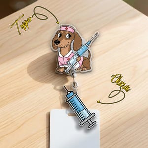 May include: A clear badge reel with a cartoon dachshund wearing a pink nurse's uniform and holding a syringe. The badge reel also features a syringe charm and a white name tag. The word "Topper" and "Charm" are written in cursive.