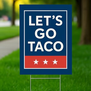 May include: A rectangular yard sign with the text "LET'S GO TACO" in white letters on a navy blue background. The bottom of the sign has a red stripe with three white stars. The sign is on a metal stake, set in green grass.