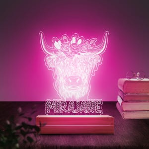 May include: A white, line art design of a Highland cow with a floral crown, illuminated in pink light. The name "MIRAJANE" is written below the cow in a decorative font. The light is on a wooden base.