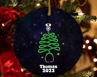 May include: A round Christmas ornament with a dark blue background and a green Christmas tree design made of a stethoscope. The ornament features a nurse's hat and the name "Thomas" with the year "2023" in white text.