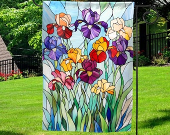 May include: A garden flag featuring a stained-glass design of colorful iris flowers. The irises are in shades of red, purple, yellow, and pink, with green leaves and a light blue background. The flag is hanging on a black metal stand in a grassy yard.
