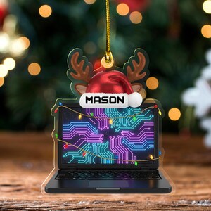 May include: A Christmas ornament shaped like a laptop with a circuit board design on the screen. The ornament features a red Santa hat with the name "MASON" and reindeer antlers. A string of colorful lights is wrapped around the laptop.