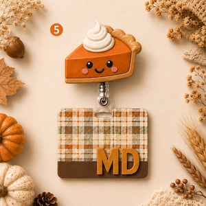 May include: A badge reel featuring a smiling pumpkin pie slice with whipped cream topping attached to a rectangular ID holder. The holder has a tan, green, and orange plaid pattern with a solid brown bottom section and the letters MD in gold.