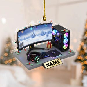 May include: A gaming-themed Christmas ornament featuring a computer setup with a curved monitor, keyboard, mouse, and headset. The monitor displays a winter scene, and the setup is decorated with Christmas lights and holly. A gold nameplate is at the bottom.