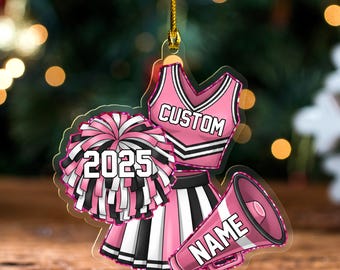 May include: A pink and white cheerleader ornament with a pom-pom, skirt, top, and megaphone. The ornament has the text "CUSTOM", "NAME", and "2025". The ornament is hanging from a gold string.