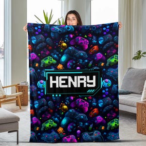 May include: A black fleece blanket with a colorful gamer controller pattern and the name "HENAY" in a blue and white box.