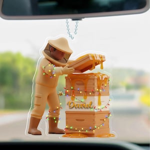 May include: A car hanging ornament featuring a beekeeper in a protective suit, opening a beehive. The beehive is stacked with honey dripping down, and the name "David" is written on the side. The ornament is decorated with colorful string lights.