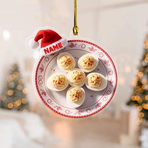 May include: A Christmas ornament featuring a plate of deviled eggs with paprika, and a Santa hat with the name "NAME". The plate has a red and white floral design. The ornament hangs from a gold string.