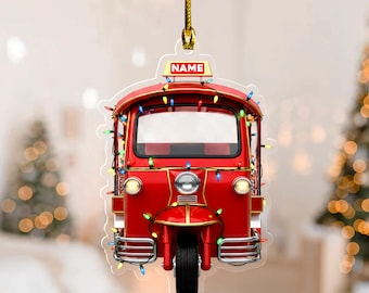 May include: A red tuk-tuk ornament with a gold hanging string. The ornament is decorated with colorful string lights and has the word "NAME" on a sign. The tuk-tuk has a single front wheel and two headlights.