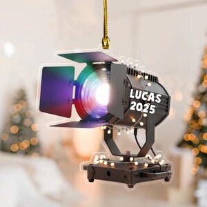 May include: A decorative ornament shaped like a stage spotlight. The spotlight is black with a bright, colorful light emanating from the lens. The text "LUCAS 2025" is printed on the side. The ornament is trimmed with small white lights and has a gold hanging cord.