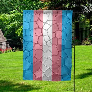 May include: A garden flag with a transgender pride flag design. The flag is made of a blue, pink, and white fabric with a textured pattern. The flag is hanging on a black metal pole.