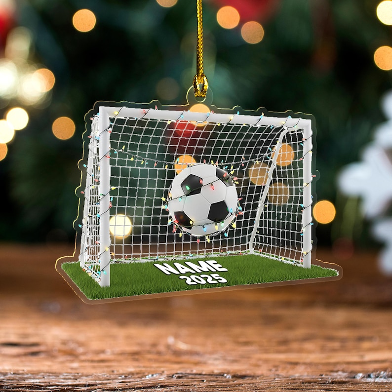 May include: A clear acrylic soccer ornament featuring a soccer ball in a net, with the text "NAME 2025" on the base. The ornament is decorated with colorful lights and a gold string for hanging.