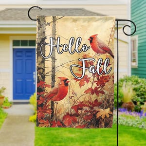 May include: A garden flag featuring a watercolor design with two red cardinal birds perched among autumn leaves. The flag has the words "Hello Fall" in a script font. The flag is displayed on a black metal stand.