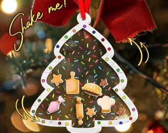 May include: A Christmas tree-shaped ornament with a clear center filled with colorful sprinkles and baking-themed charms. The ornament has a white border and a red ribbon for hanging. Text on the ornament says "Shake me!"