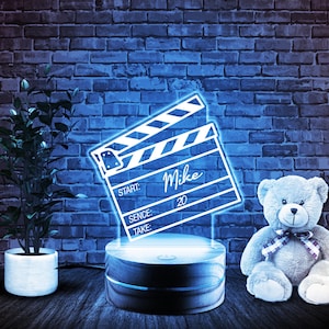 May include: A blue neon light shaped like a clapperboard with the name "Mike" and the number "20" written on it. The light is sitting on a wooden base. A gray teddy bear is sitting in front of the light.