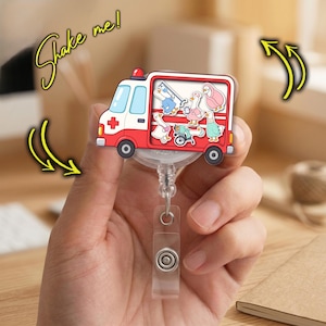 May include: A red and white ambulance-shaped badge reel featuring a clear window filled with small, loose cartoon goose characters dressed as medical staff. The text "Shake me!" is written in yellow script with arrows indicating movement.