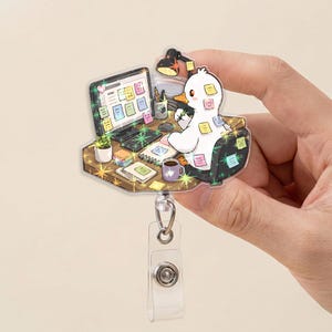 May include: An acrylic badge reel featuring a white duck sitting at a desk with a computer, a lamp, a potted plant, and a coffee mug. The duck is covered in colorful sticky notes, and the entire scene is decorated with sparkling star accents.