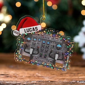 May include: A Christmas ornament shaped like an airplane cockpit, with the name "LUCAS" and the year "2025" on a Santa hat. The ornament is decorated with colorful lights and has a gold string for hanging.