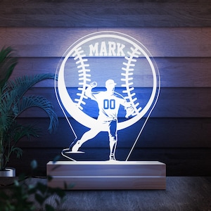 May include: A baseball-themed LED lamp featuring a baseball player silhouette throwing a ball, set against a baseball design. The name "MARK" is displayed at the top. The lamp is mounted on a wooden base, creating a decorative and illuminated piece.