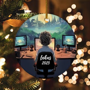 May include: A round Christmas ornament featuring a digital illustration of a person sitting in front of two computer monitors with a cityscape in the background. The person is wearing headphones and the text "Lukas 2023" is on the chair.