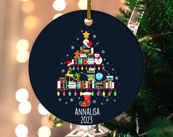 May include: A navy blue Christmas ornament with a gold string. The ornament features a Christmas tree design made of books, a star, and other festive elements. The name "ANNALISA" and the year "2023" are printed on the ornament.