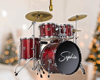 May include: A hanging ornament shaped like a metallic red drum set with silver hardware, gold-colored cymbals, and a black bass drum head featuring the white script name Sophia. The ornament is suspended by a thin gold cord for display.