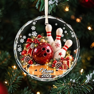 May include: A round, faceted glass Christmas ornament featuring a bowling theme. The design includes a red bowling ball, three white bowling pins, pine branches, red berries, pinecones, and gift boxes. The text "Name" and "202x" is printed below.