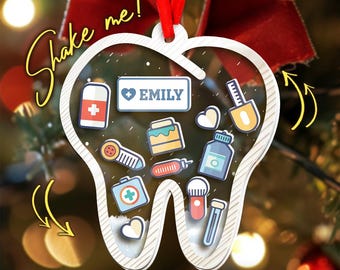 May include: A white tooth-shaped Christmas ornament with a red ribbon. Inside the ornament are various medical-themed icons and the name "EMILY". The text "Shake me!" is written in a cursive font.