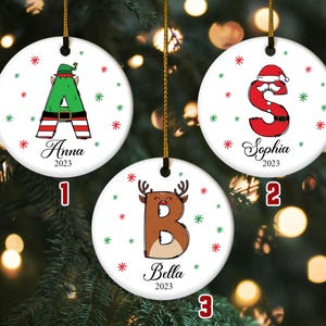 May include: Three white ceramic Christmas ornaments with gold string. Each ornament features a letter, name, and year. The letters are decorated with festive designs: an elf, Santa, and a reindeer. The names are Anna, Sophia, and Bella.
