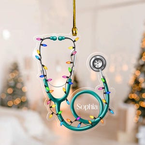 May include: A teal stethoscope Christmas ornament with colorful lights. The ornament has the name "Sophia" on it and is hanging from a gold string. The background is blurred with Christmas trees.